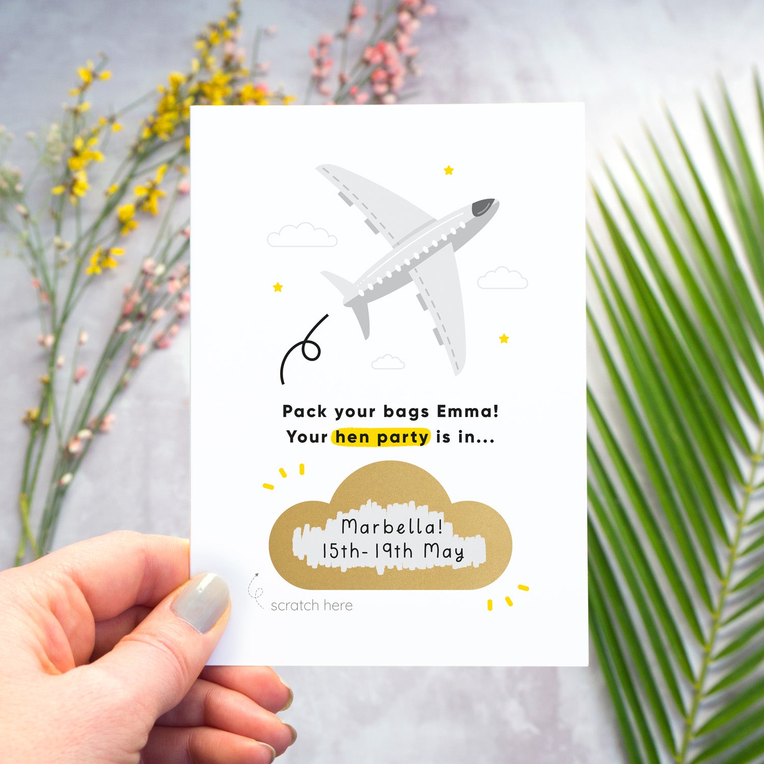 A hen party scratch and reveal card featuring a grey aeroplane and scratch off gold cloud that has been photographed being held over a grey background with yellow and pink flowers and green foliage. The cloud has been scratched off to reveal the hen do destination and date.