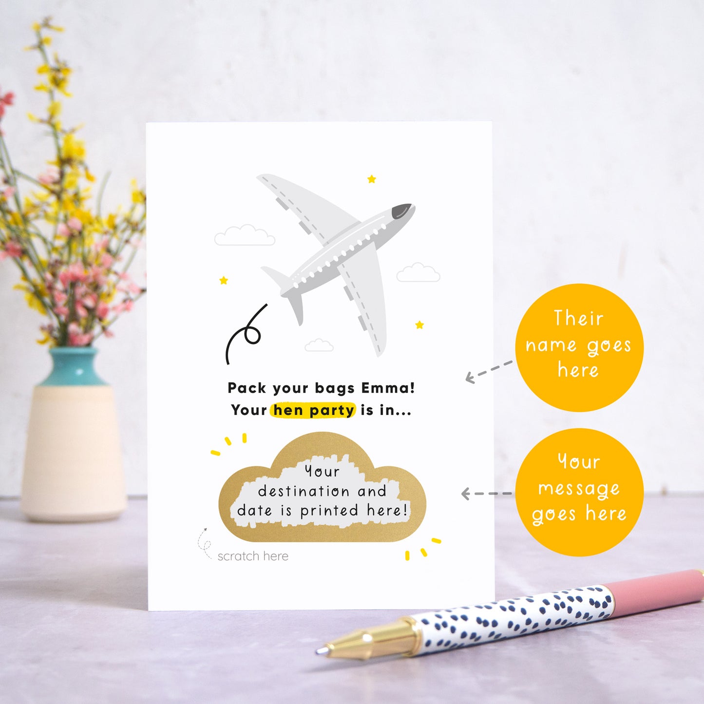 A hen party scratch and reveal card featuring a grey aeroplane and scratch off gold cloud that has been photographed stood up on a grey background with a small jar of flowers and a pen for scale. The card is surrounded by orange circles which point to the areas that can be personalised. These areas are the name and the message for under the scratch off panel.