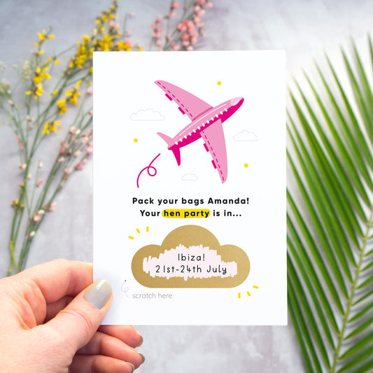 A hen party scratch and reveal card featuring a pink aeroplane and scratch off gold cloud that has been photographed being held over a grey background with yellow and pink flowers and green foliage. The cloud has been scratched off to reveal the hen do destination and date.