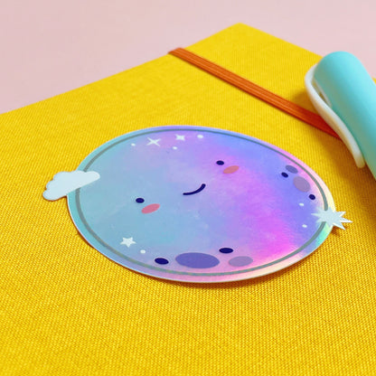 A shiny iridescent holographic vinyl sticker featuring a smiling moon lying on top of a yellow journal on a pink background