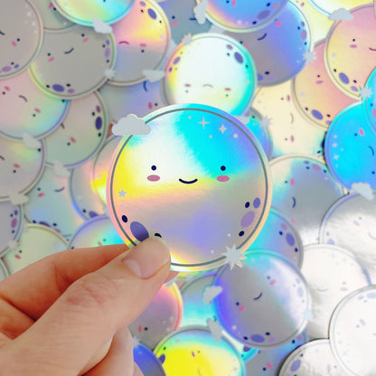 A shiny iridescent holographic vinyl sticker featuring a smiling moon being held over a pile of stickers of the same design