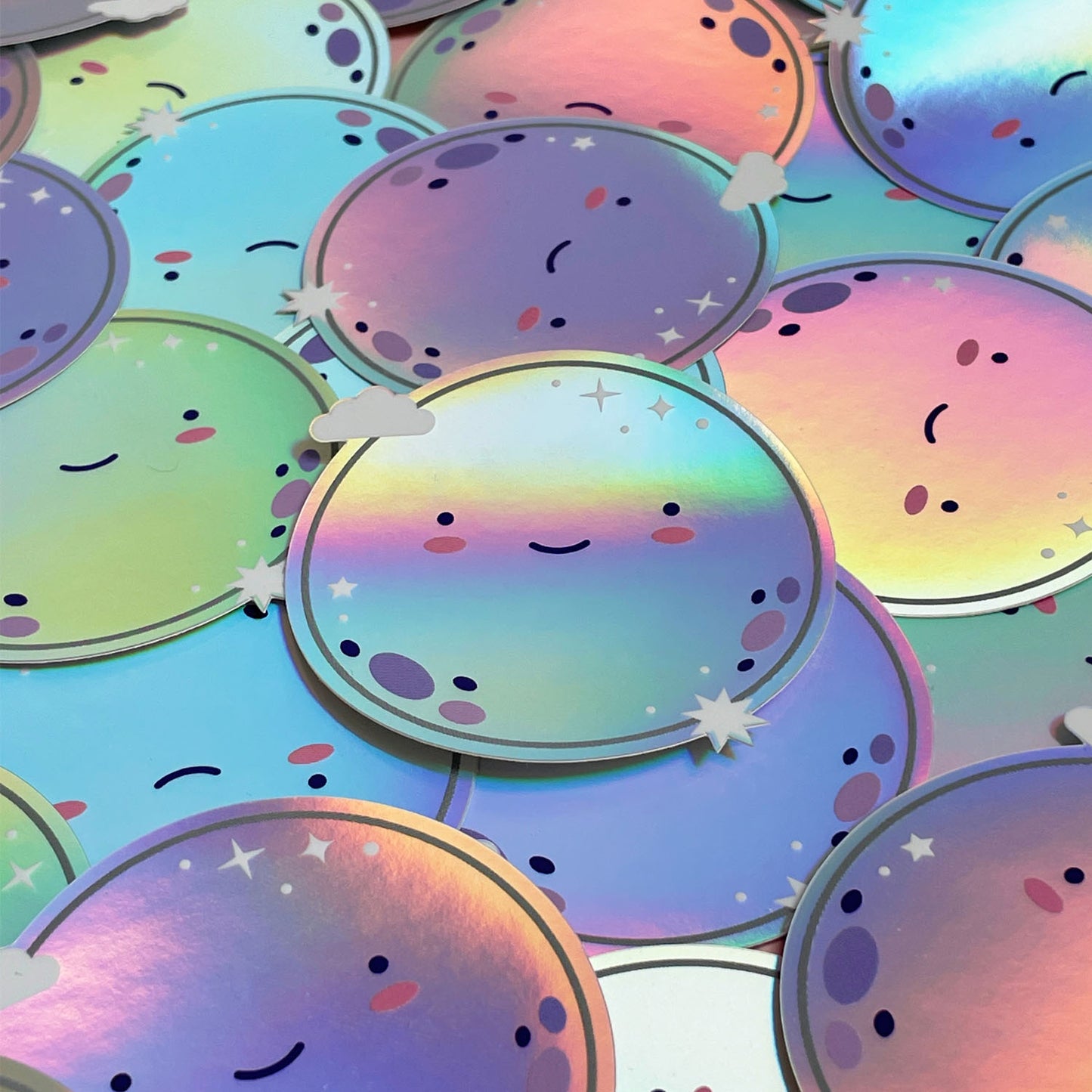 A pile of shiny iridescent holographic vinyl sticker featuring a smiling moon. The surface is covered with dozens of the same design.