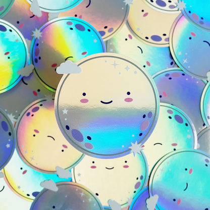 A pile of shiny iridescent holographic vinyl sticker featuring a smiling moon. The surface is covered with dozens of the same design.