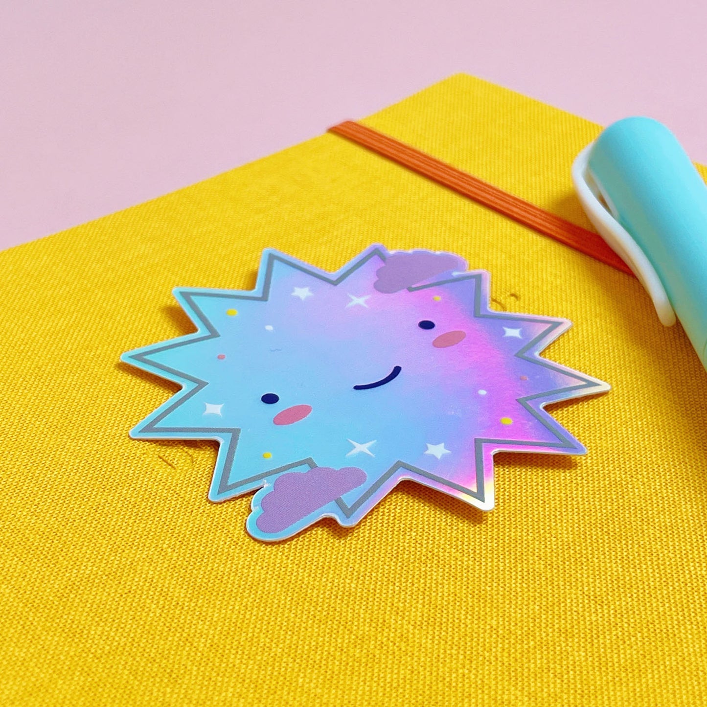 A shiny iridescent holographic vinyl sticker featuring a smiling starburst lying on top of a yellow journal on a pink background