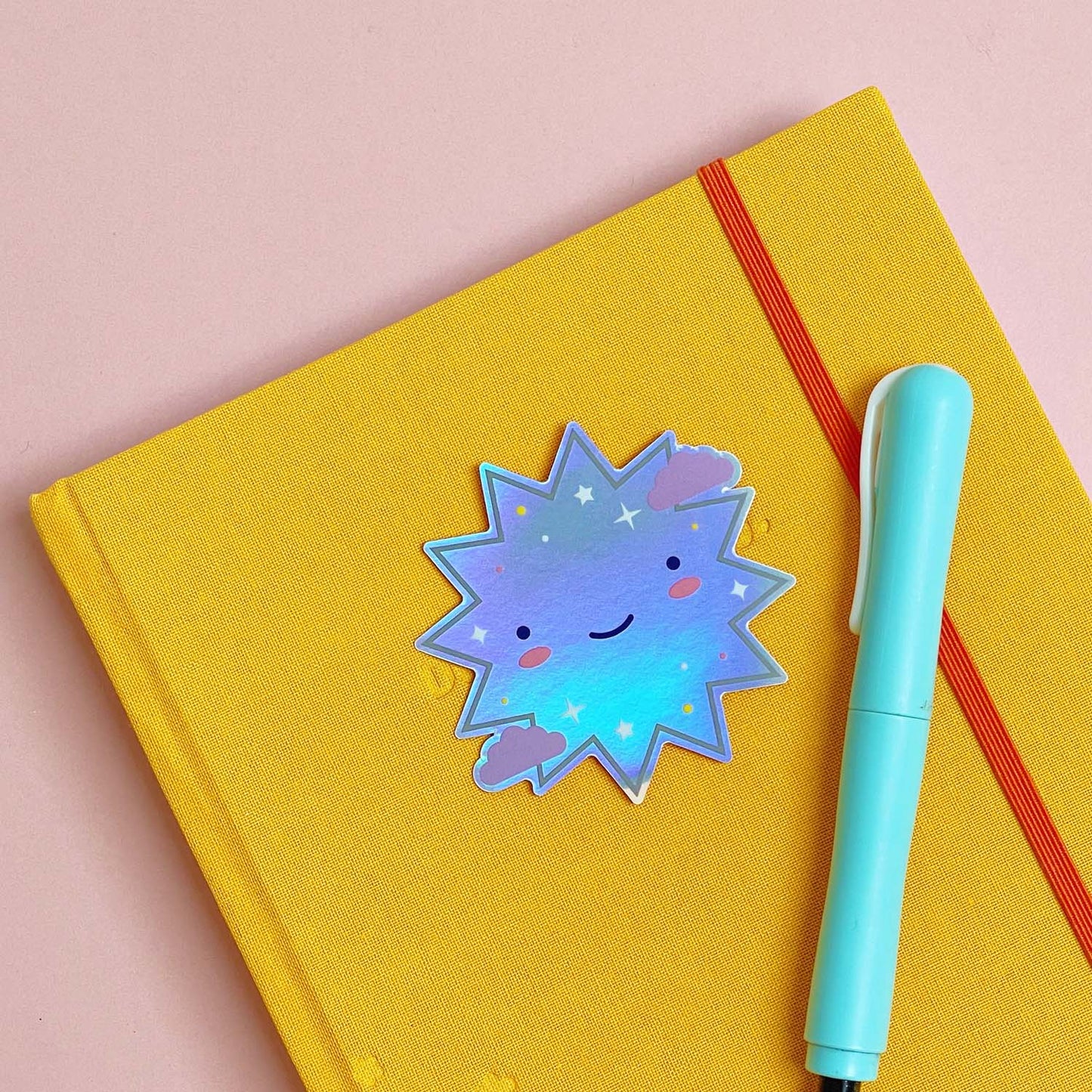 A shiny iridescent holographic vinyl sticker featuring a smiling starburst lying on top of a yellow journal on a pink background
