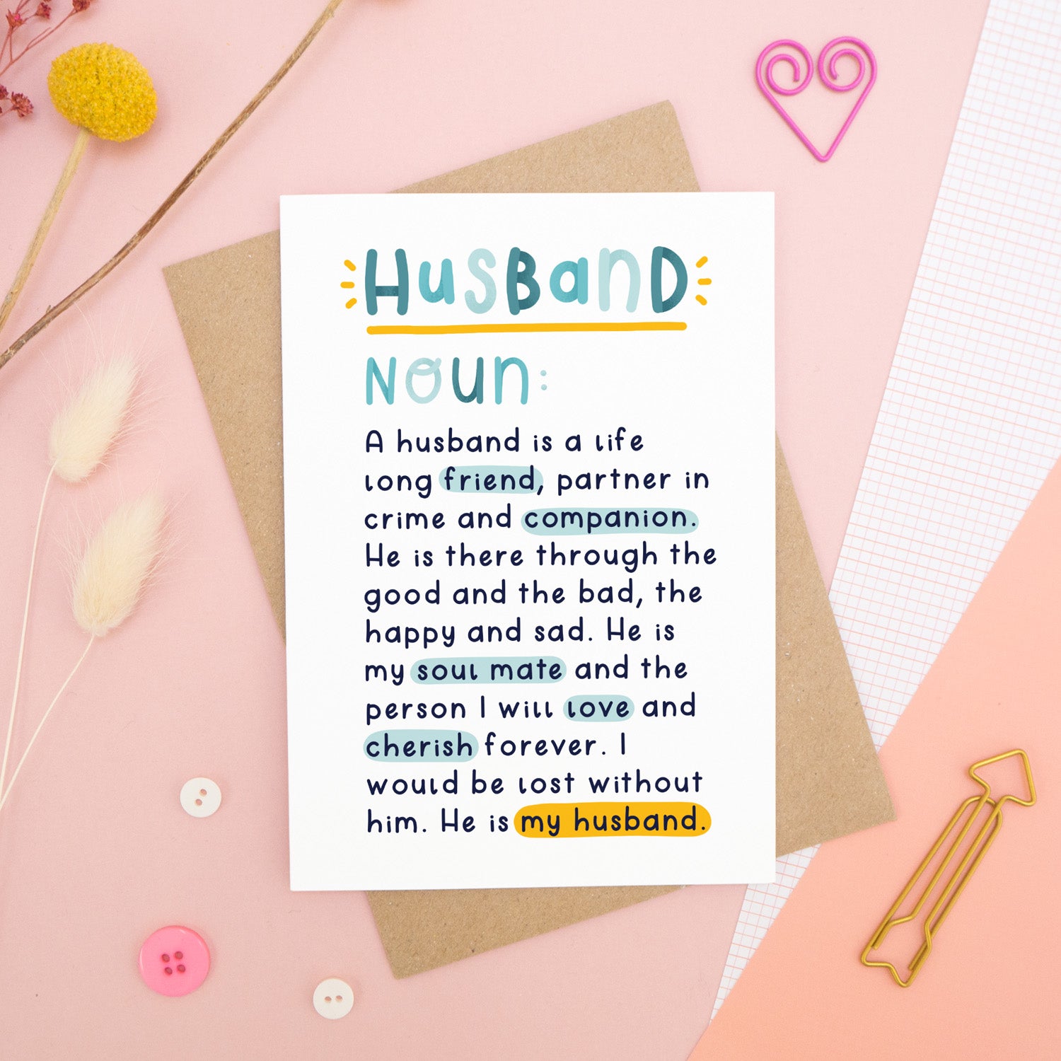 A heartfelt, colourful dictionary definition style card with the definition of a husband, photographed on a pink background with floral props, paper clips, and buttons. 