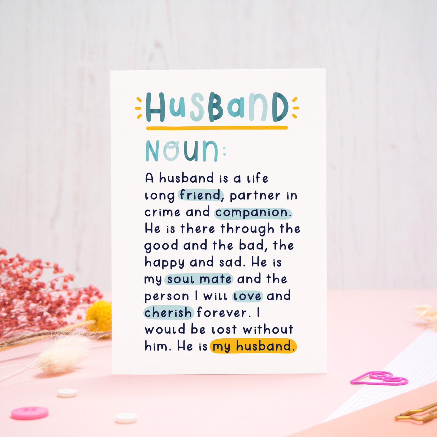 A heartfelt, colourful dictionary definition style card with the definition of a husband, photographed stood on a pink and white background with floral props, paper clips, and buttons. 