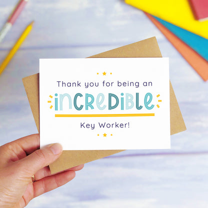 An incredible key worker nursery thank you card being held over a blue background with colourful text books and two pens. The card is sat on top of its kraft brown envelope. This is the teacher card with the blue text colour option.