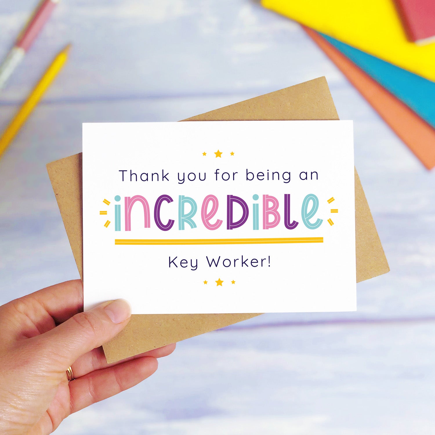 An incredible key worker nursery thank you card being held over a blue background with colourful text books and two pens. The card is sat on top of its kraft brown envelope. This is the teacher card with the pink, purple and blue text colour option.