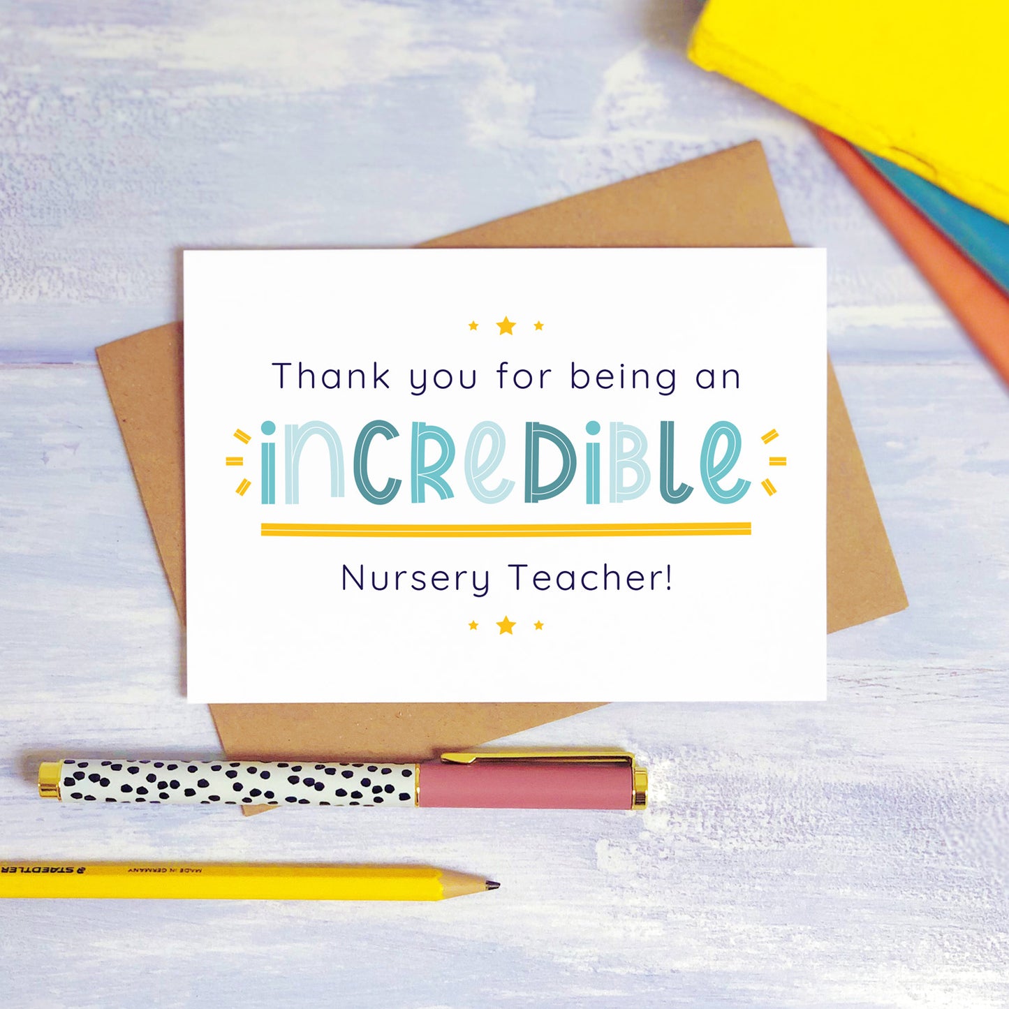 An incredible nursery teacher thank you card lying on top of a kraft brown envelope, flat lay style on a blue textured background with colourful text books, a pen and a pencil. This nursery teacher card features the blue text colour option.