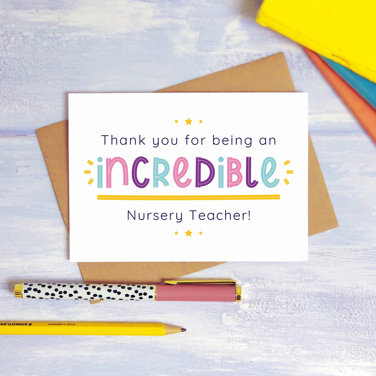 An incredible nursery teacher thank you card lying on top of a kraft brown envelope, flat lay style on a blue textured background with colourful text books, a pen and a pencil. This nursery teacher card features the pink, purple and blue text colour option.