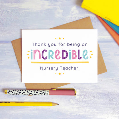 An incredible nursery teacher thank you card lying on top of a kraft brown envelope, flat lay style on a blue textured background with colourful text books, a pen and a pencil. This nursery teacher card features the pink, purple and blue text colour option.