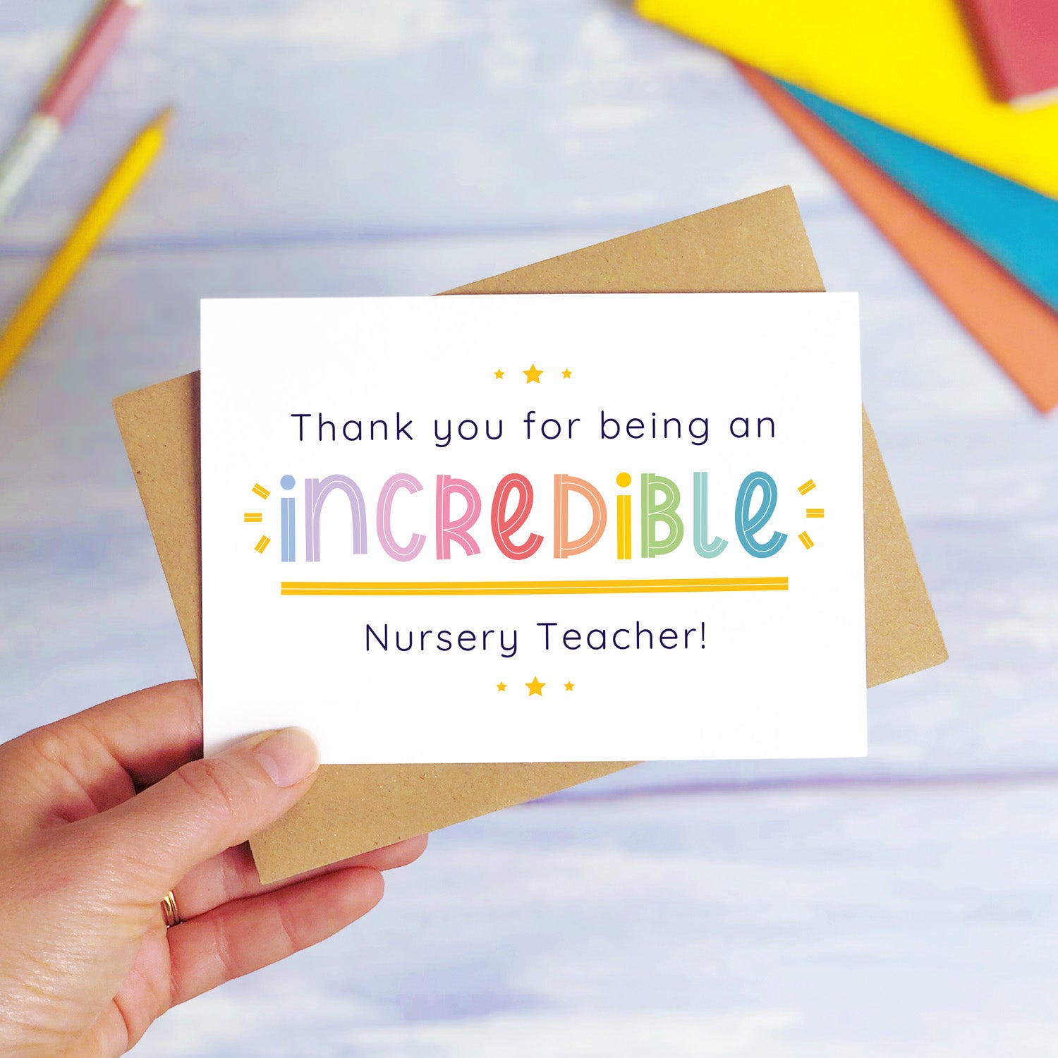 An incredible nursery teacher thank you card being held over a blue background with colourful text books and two pens. The card is sat on top of its kraft brown envelope. This is the nursery teacher card with the rainbow text colour option.