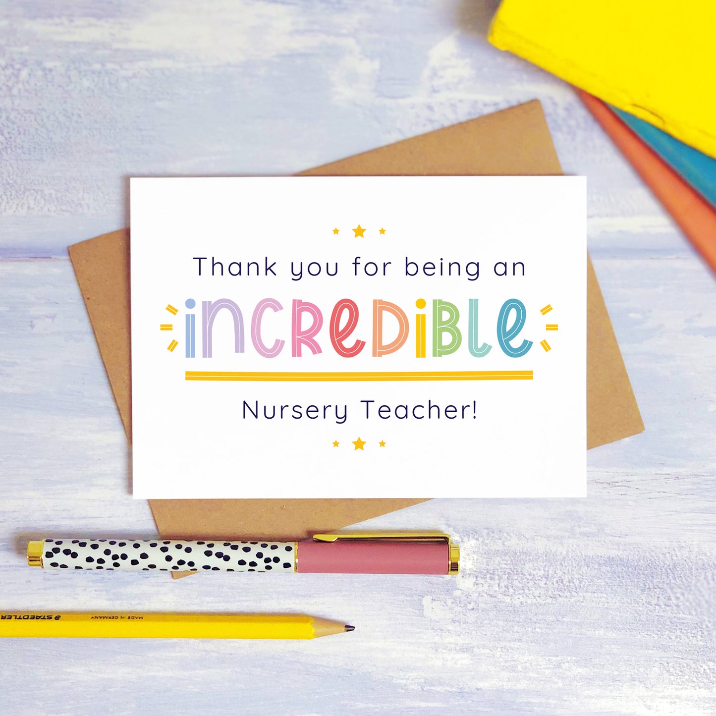 An incredible nursery teacher thank you card lying on top of a kraft brown envelope, flat lay style on a blue textured background with colourful text books, a pen and a pencil. This nursery teacher card features the rainbow text colour option.