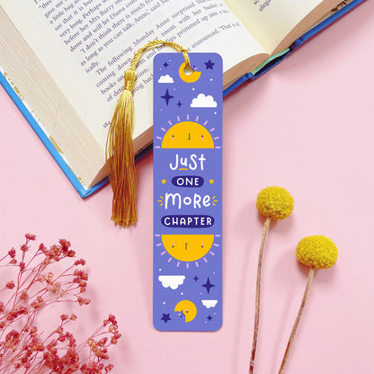 A celestial themed, double sided, metal bookmark with a gold tassel which has been photographed on a pink background with an open book and dried flowers.