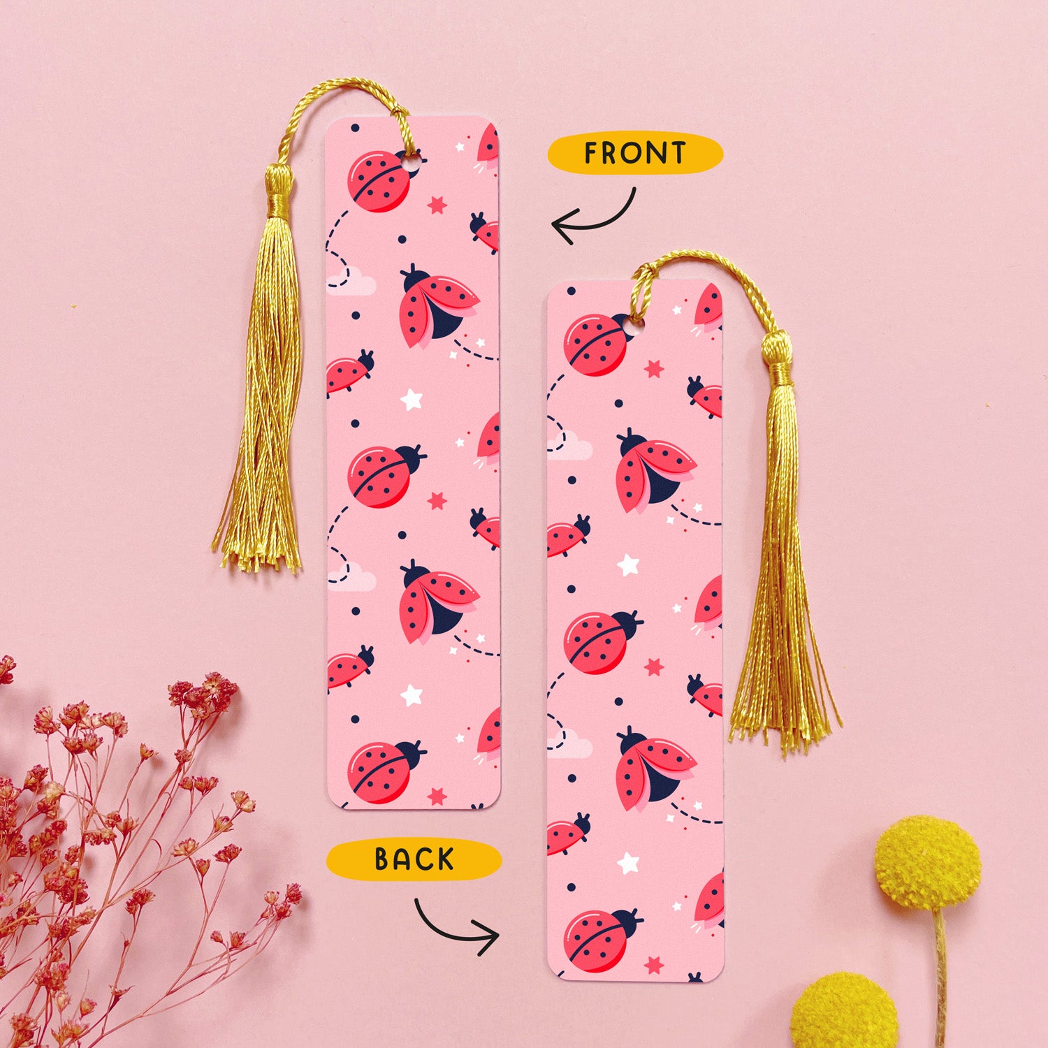 An image depicting the front and the back of the double sided metal bookmark which has been photographed on a pink background with dried flowers. Each bookmark has a gold tassel. The front and the back of the bookmark are printed with the same design of a ladybugs, stars and clouds on a pink sky.