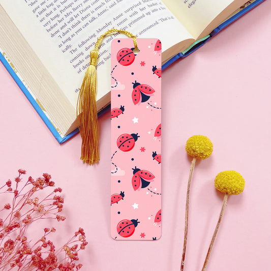 A ladybug themed, double sided, metal bookmark with a gold tassel which has been photographed on a pink background with an open book and dried flowers.