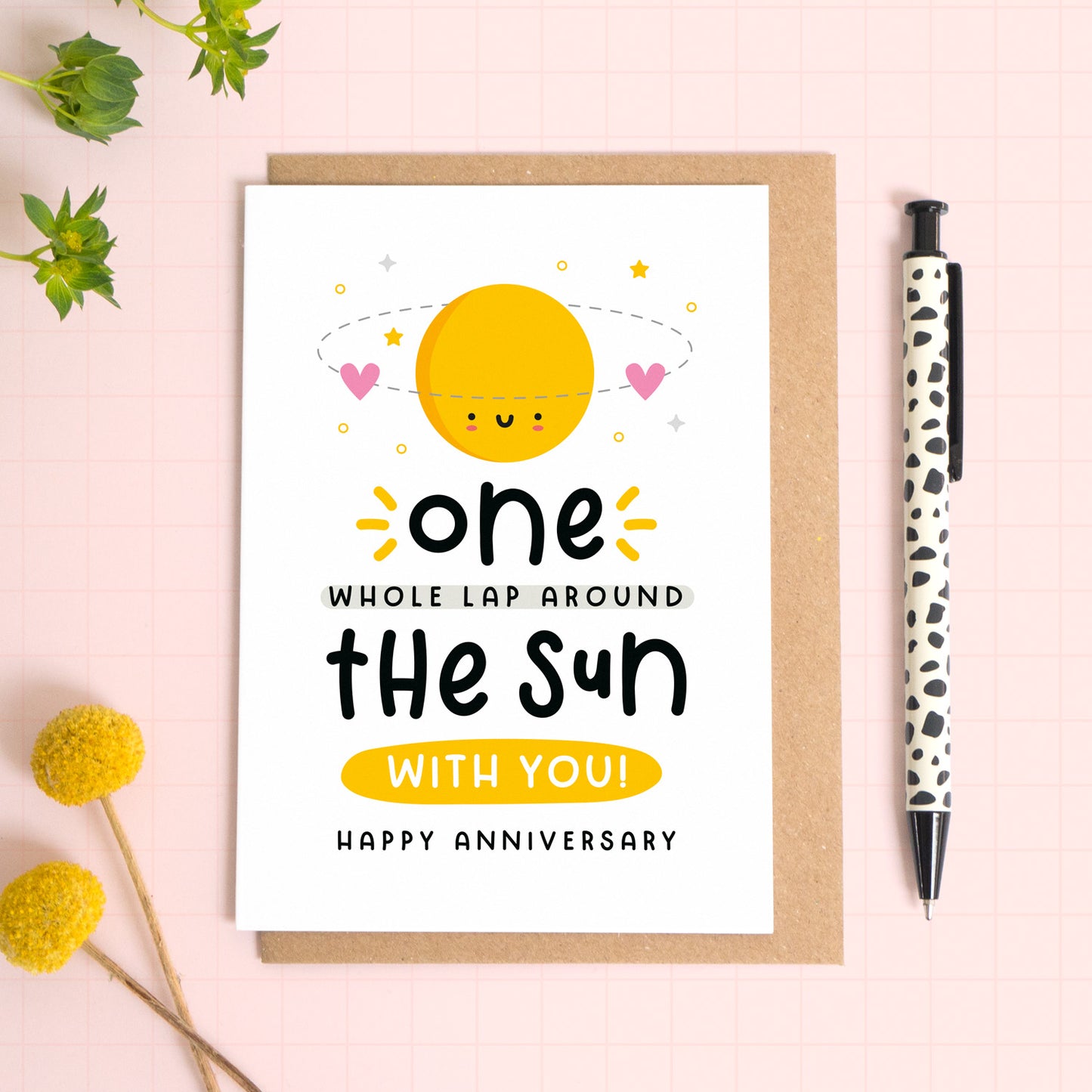 A 1 lap around the sun wedding anniversary card for your 1st wedding anniversary which has been photographed on a Kraft brown envelope upon a pink background surrounded by florals and a pen for scale.