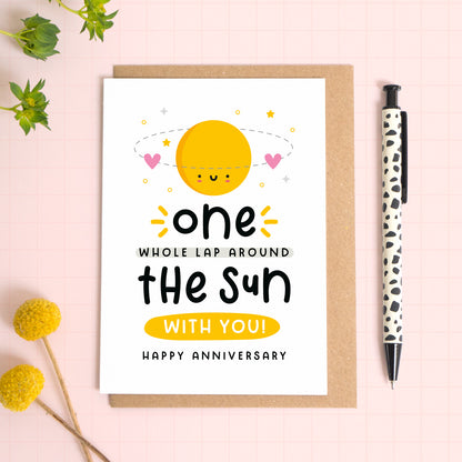 A 1 lap around the sun wedding anniversary card for your 1st wedding anniversary which has been photographed on a Kraft brown envelope upon a pink background surrounded by florals and a pen for scale.