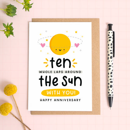 A 10 laps around the sun wedding anniversary card for your 10th wedding anniversary which has been photographed on a Kraft brown envelope upon a pink background surrounded by florals and a pen for scale.