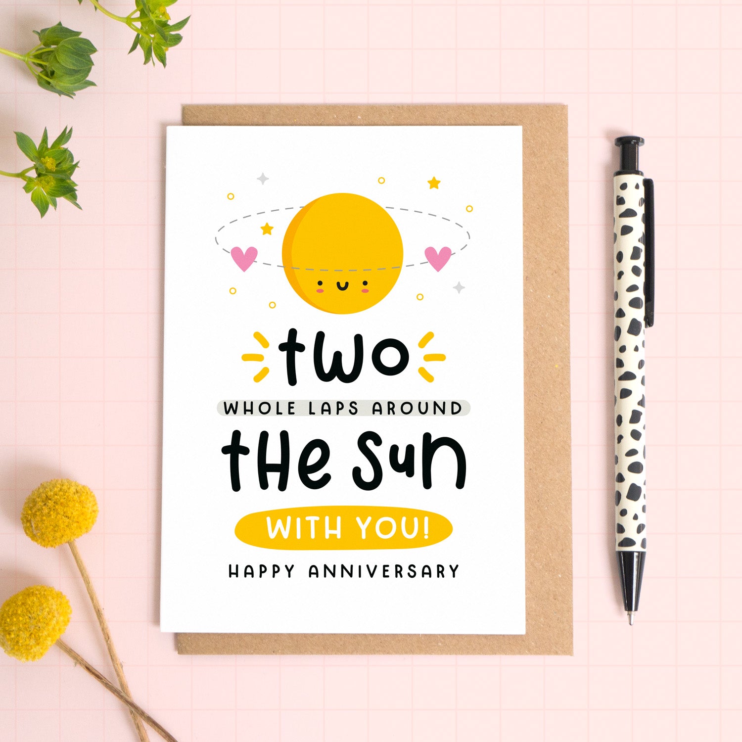 A 2 laps around the sun wedding anniversary card for your 2nd wedding anniversary which has been photographed on a Kraft brown envelope upon a pink background surrounded by florals and a pen for scale.