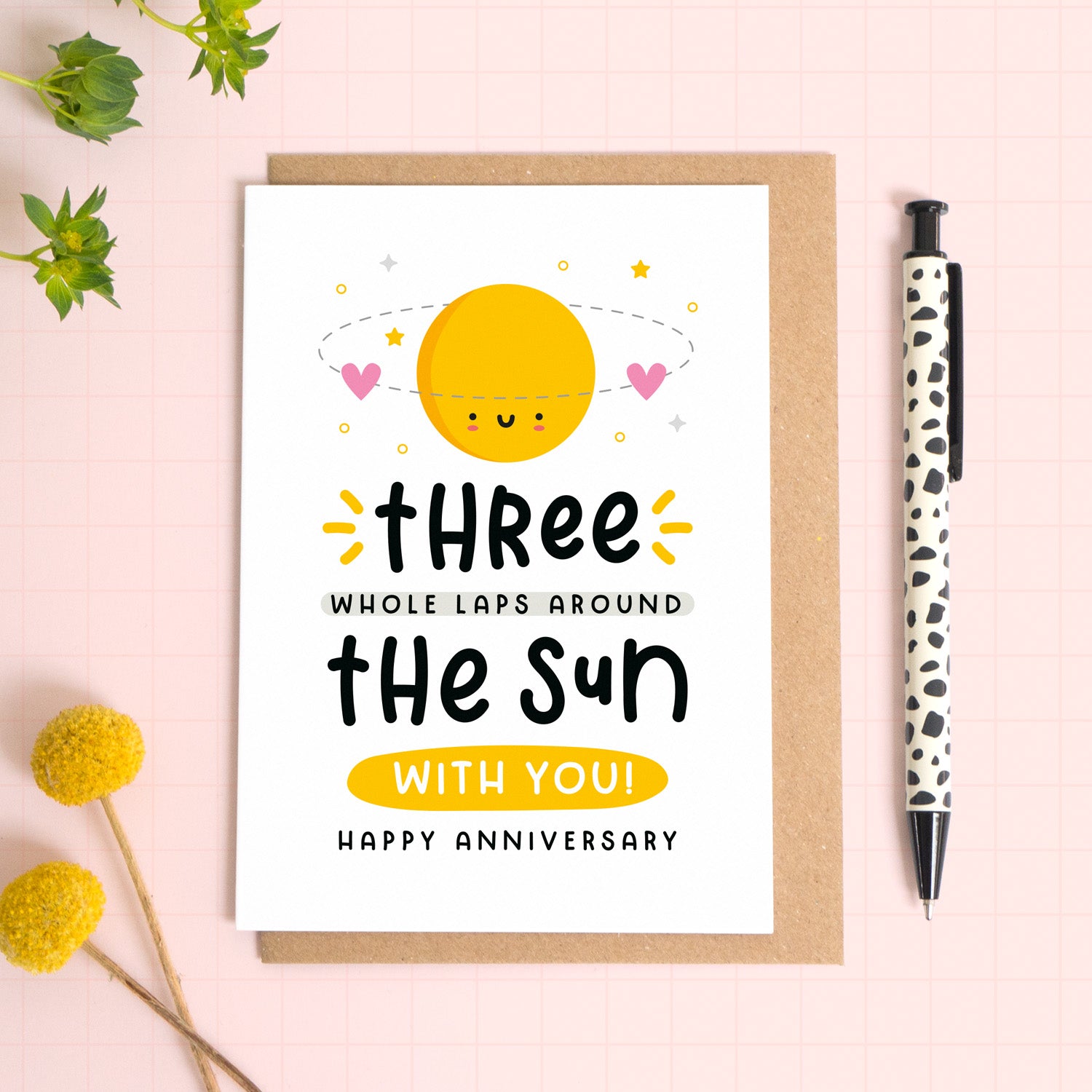 A 3 laps around the sun wedding anniversary card for your 3rd wedding anniversary which has been photographed on a Kraft brown envelope upon a pink background surrounded by florals and a pen for scale.