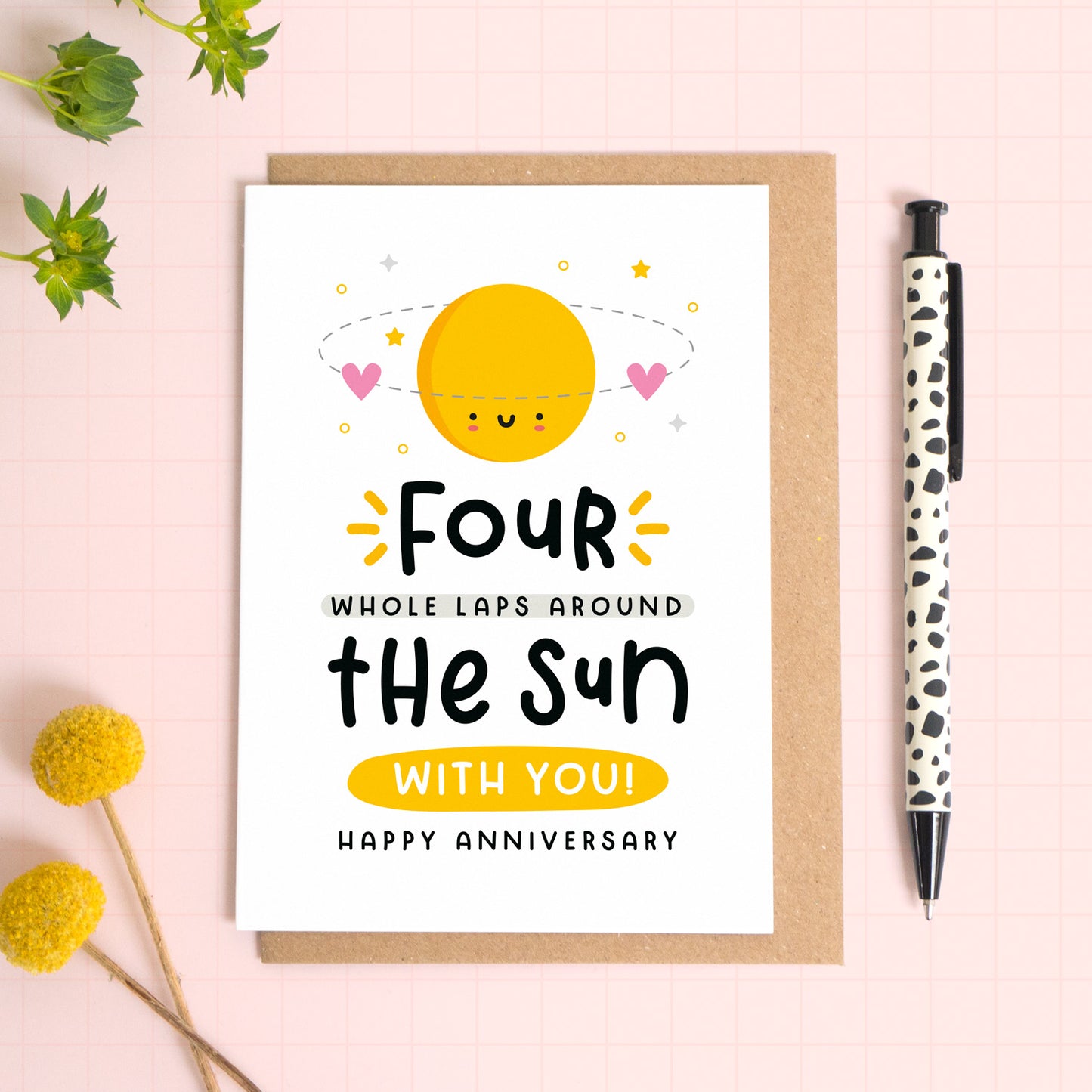 A 4 laps around the sun wedding anniversary card for your 4th wedding anniversary which has been photographed on a Kraft brown envelope upon a pink background surrounded by florals and a pen for scale.