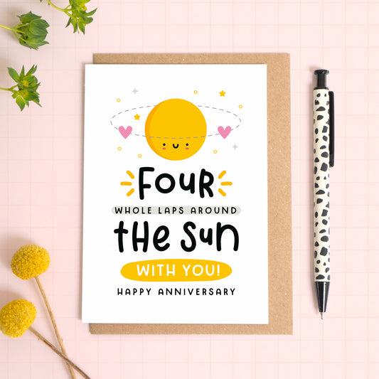 A 4 laps around the sun wedding anniversary card for your 4th wedding anniversary which has been photographed on a Kraft brown envelope upon a pink background surrounded by florals and a pen for scale.