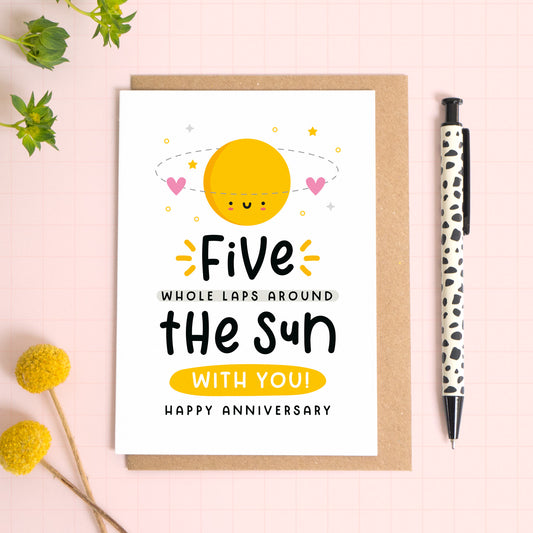 A 5 laps around the sun wedding anniversary card for your 5th wedding anniversary which has been photographed on a Kraft brown envelope upon a pink background surrounded by florals and a pen for scale.