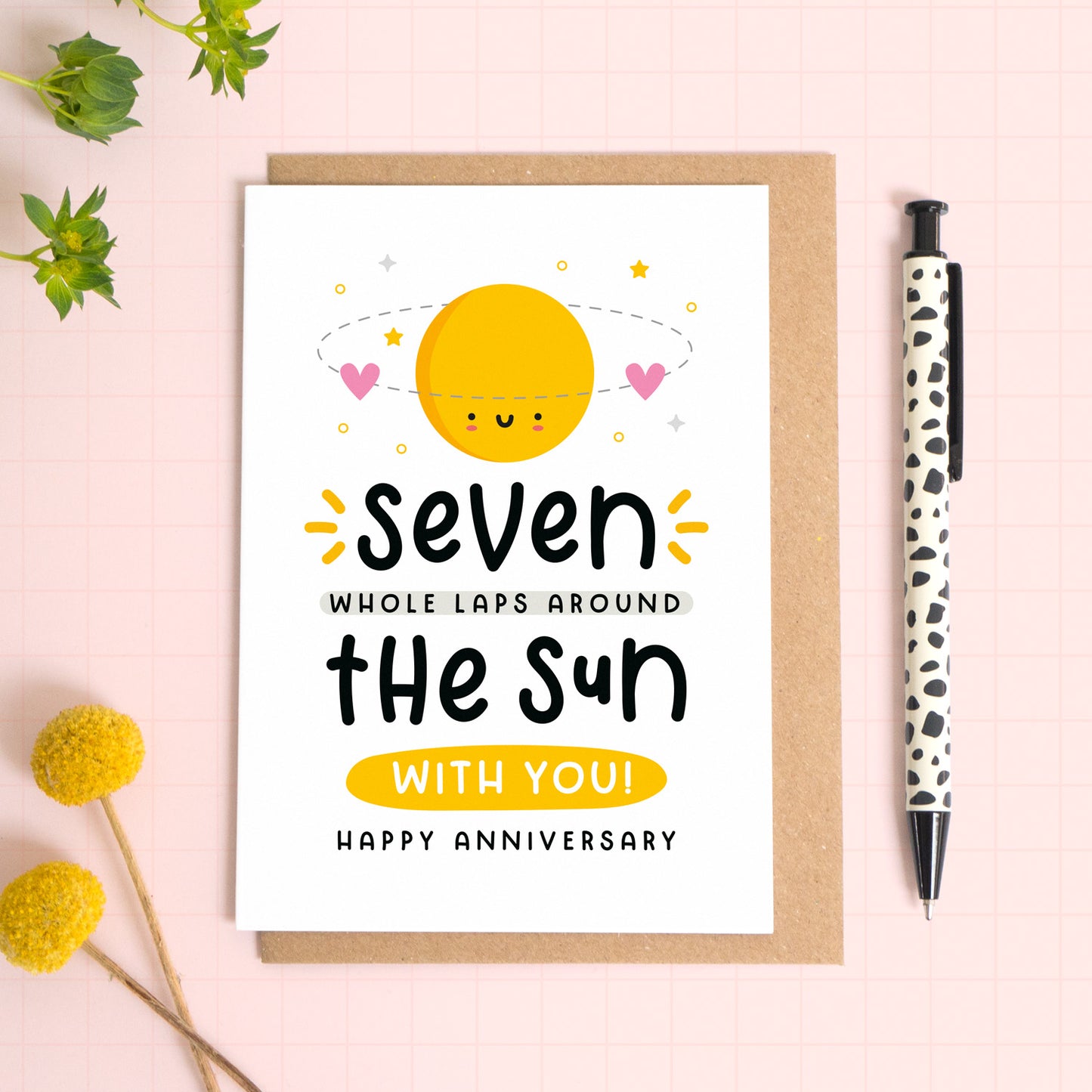 A 7 laps around the sun wedding anniversary card for your 7th wedding anniversary which has been photographed on a Kraft brown envelope upon a pink background surrounded by florals and a pen for scale.