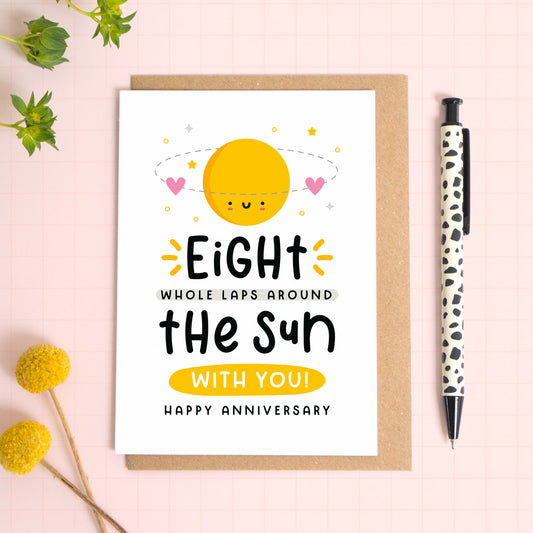 A 8 laps around the sun wedding anniversary card for your 8th wedding anniversary which has been photographed on a Kraft brown envelope upon a pink background surrounded by florals and a pen for scale.