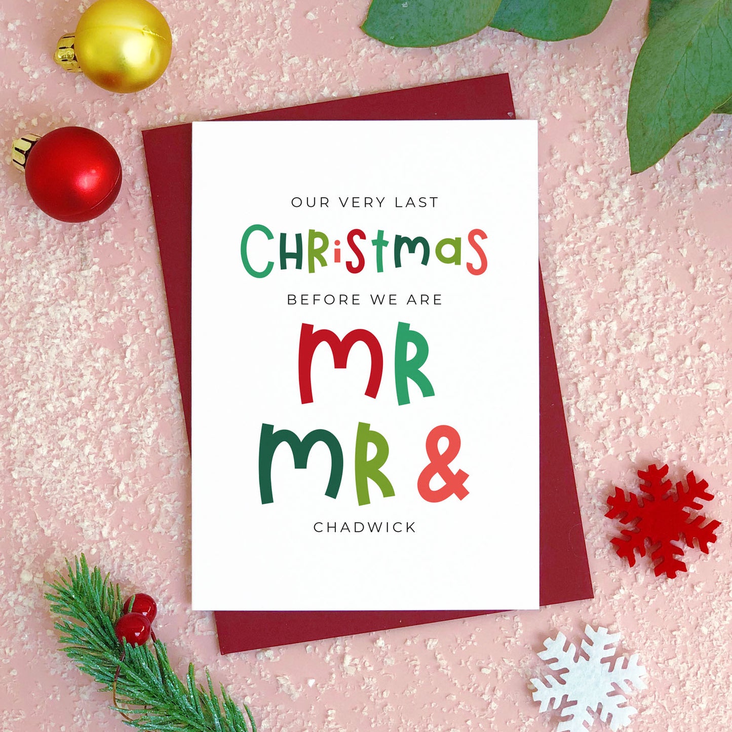 A personalised last Christmas card photographed flat lying on a red wine coloured envelope on a pink surface surrounded by fake snow, baubles and foliage.