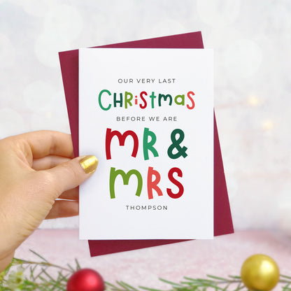 A personalised last Christmas card photographed being held by a hand to the left and foliage and baubles in the bottom of the frame. The card is against its ‘red wine’ colour envelope.