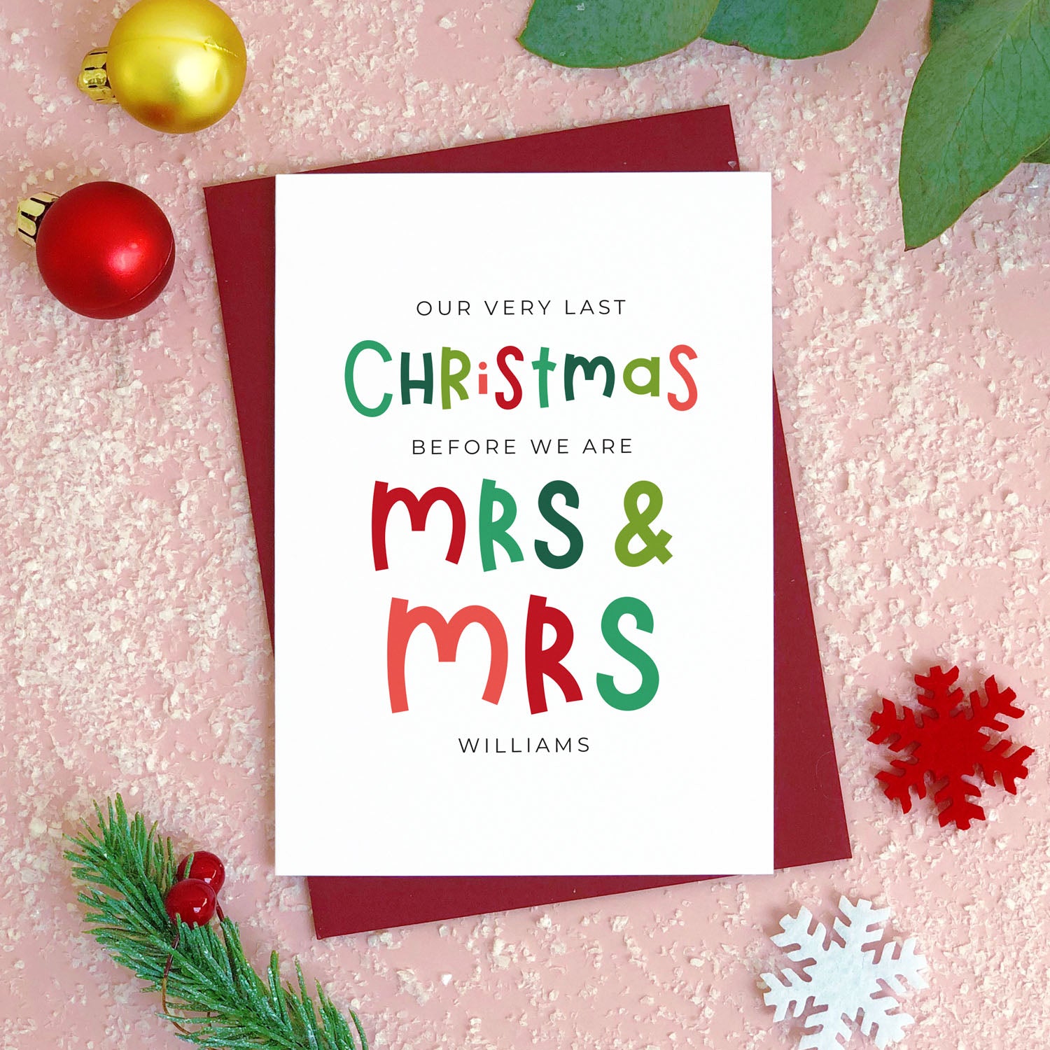 A personalised last Christmas card photographed flat lying on a red wine coloured envelope on a pink surface surrounded by fake snow, baubles and foliage.