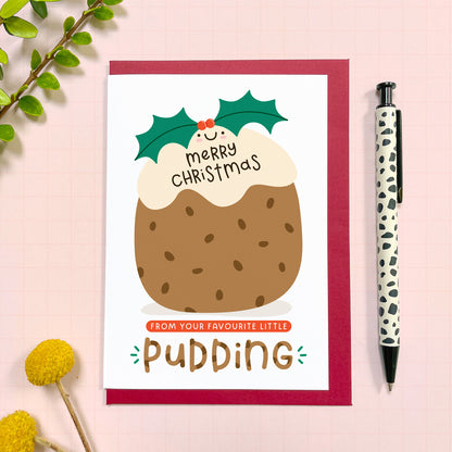 A cute Christmas pudding card from a baby, toddler or child to their parents or grandparents featuring a happy Christmas pudding. The back of the greetings card showing the Joanne Hawker logo, copyright info and website. The card has a red envelope and it has been photographed on a pink background next to yellow flowers and a pen for scale.