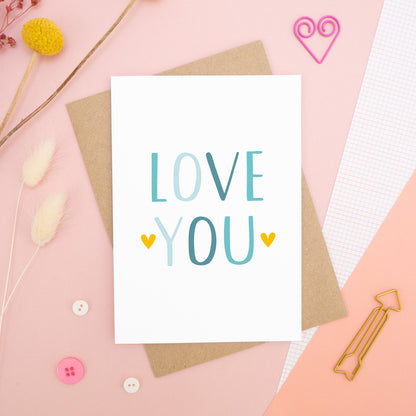 An anniversary ‘love you’ card photographed on a pink background with floral props, paper clips, and buttons. This image shows the ‘love you’ anniversary card in the blue colour way. 