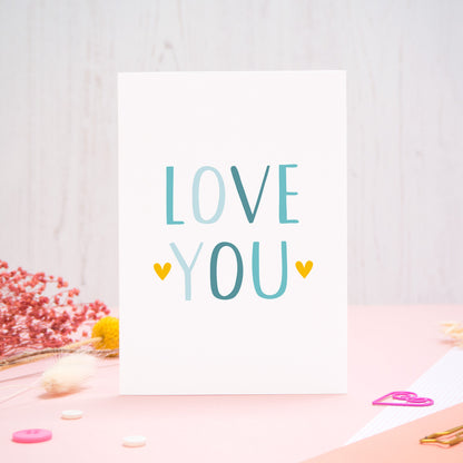 An anniversary ‘love you’ card photographed stood on a pink and white background with floral props, paper clips, and buttons. This image shows the ‘love you’ anniversary card in the blue colour way. 