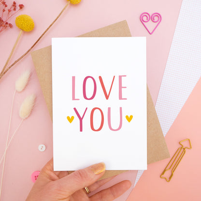 An anniversary ‘love you’ card photographed held over on a pink background with floral props, paper clips, and buttons. This image shows the ‘love you’ anniversary card in the pink colour way.
