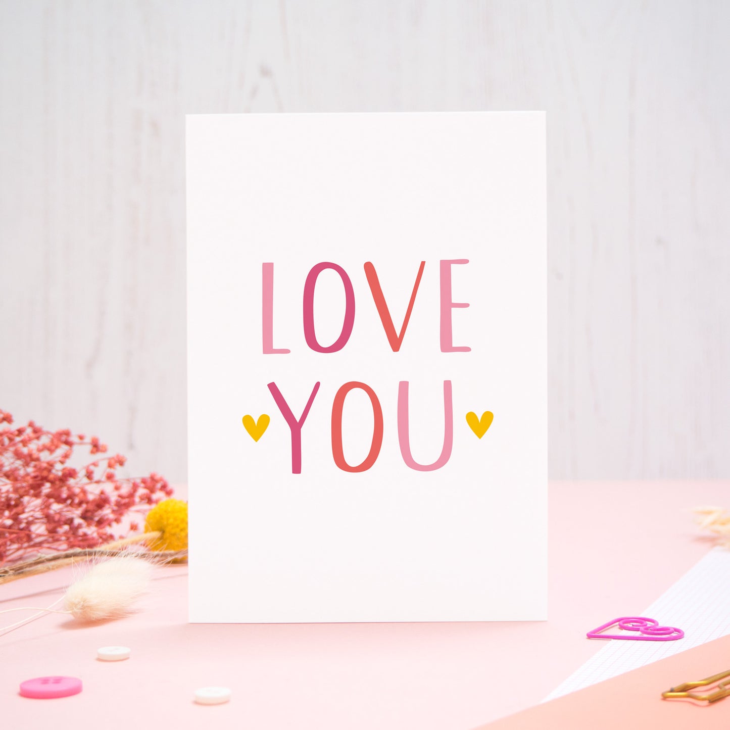 An anniversary ‘love you’ card photographed stood on a pink and white background with floral props, paper clips, and buttons. This image shows the ‘love you’ anniversary card in the pink colour way. 