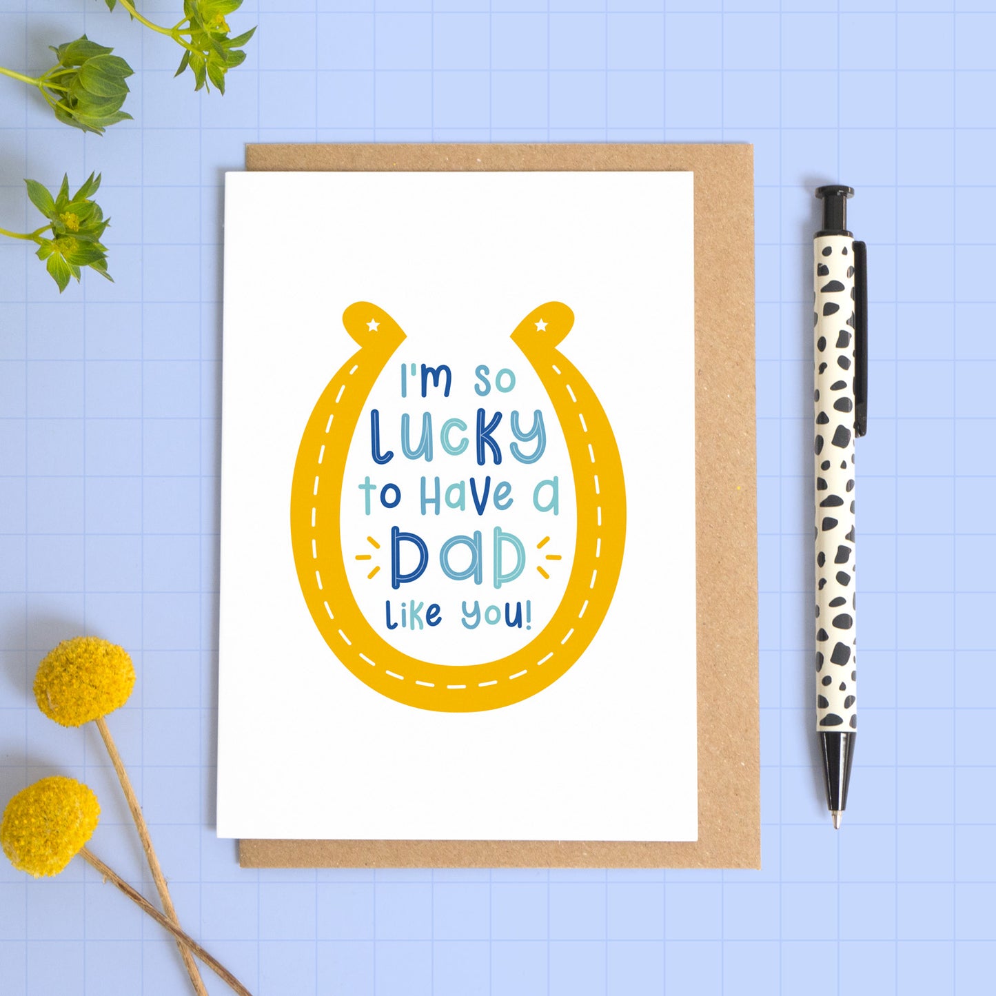 An image showing a card with writing inside of a horseshoe that reads 'I'm so lucky to have a dad like you'. The horseshoe is yellow and the text is in varying tones of blue. The card has been laid on top of a kraft brown envelope and has been photographed on a blue background. There are flowers and a pen for props.