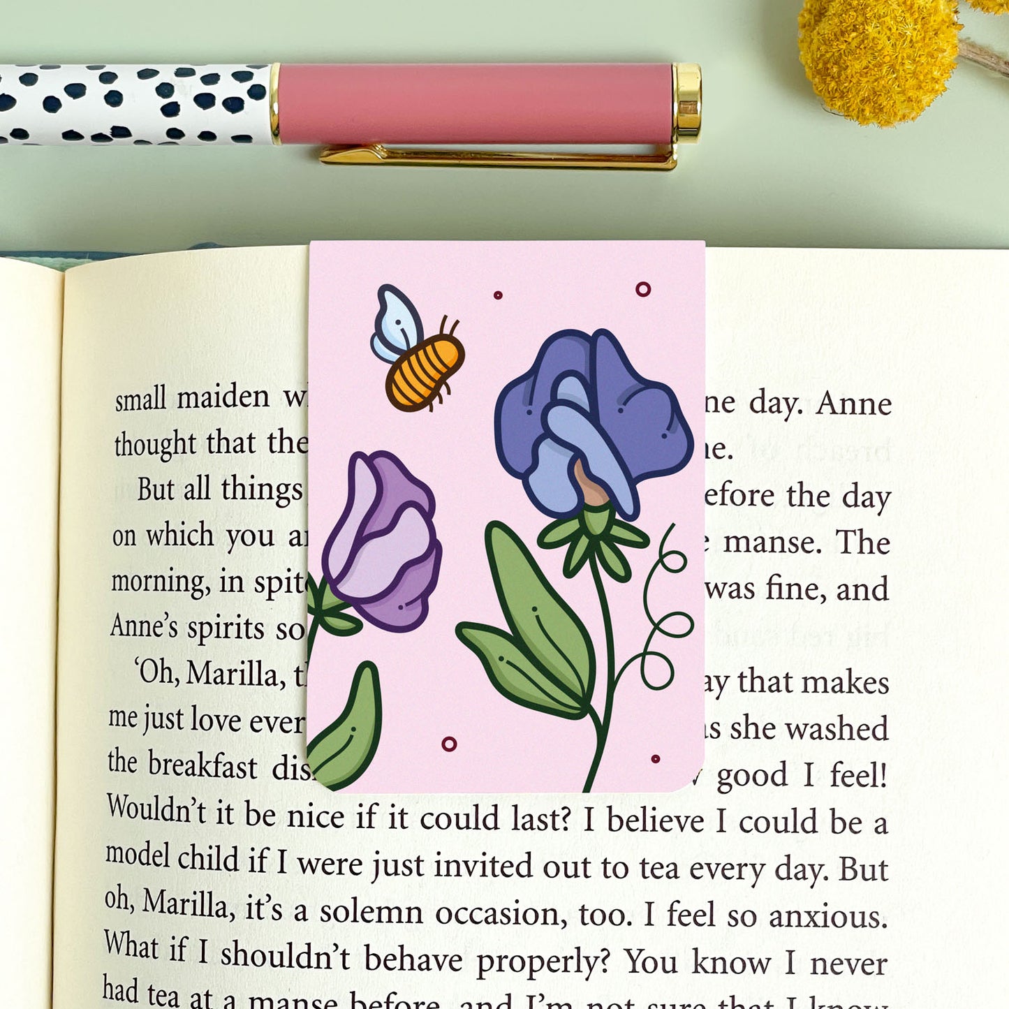 Floral blue and purple sweet pea bookmark on an open book with a polka dot pen and yellow flower in the background.