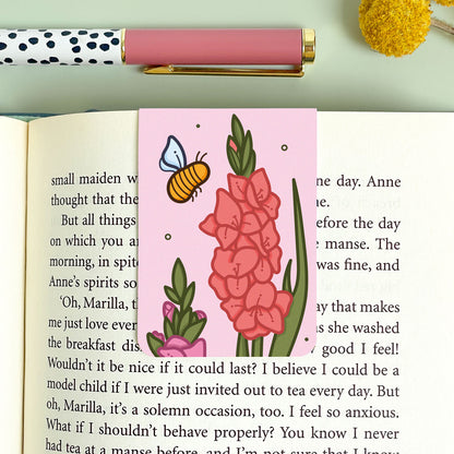 Book with a pink bookmark featuring gladiolus flowers and a bee, next to a polka dot pen on a light green background.