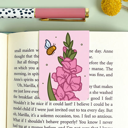 Floral gladiolus bookmark with a bee on an open book, next to a polka dot pen and yellow flower.