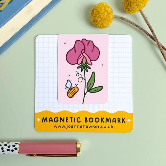 Magnetic bookmark with floral sweet pea design and bee illustration on a notebook, with a pen and decorative elements in the background.
