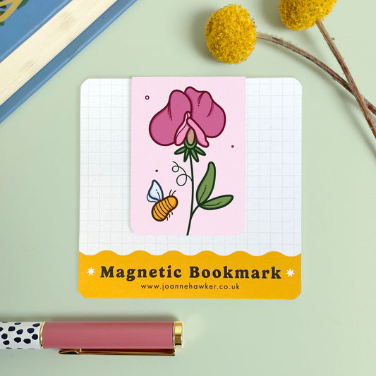 Magnetic bookmark with floral sweet pea design and bee illustration on a notebook, with a pen and decorative elements in the background.