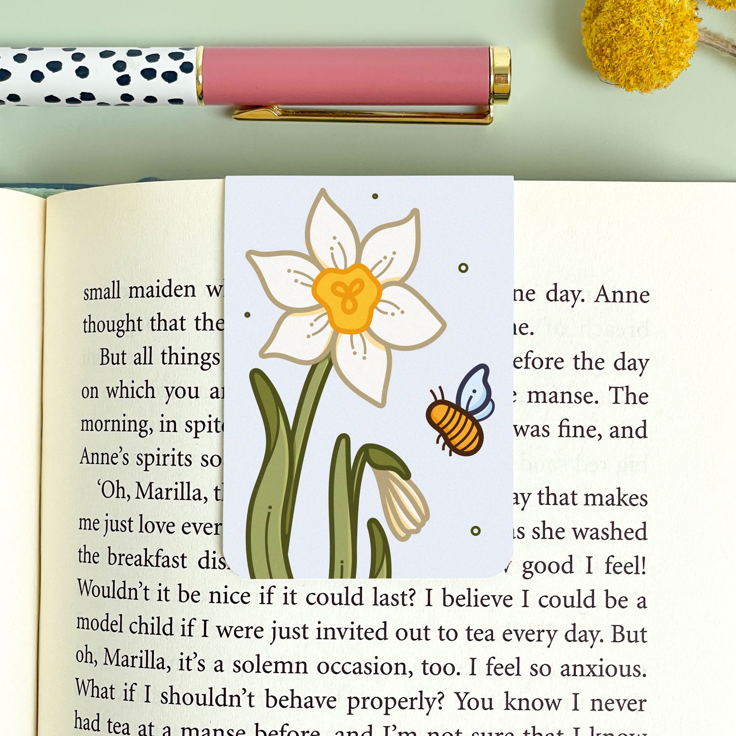 Floral narcissus bookmark with a bee on an open book, next to a pen and decorative items.