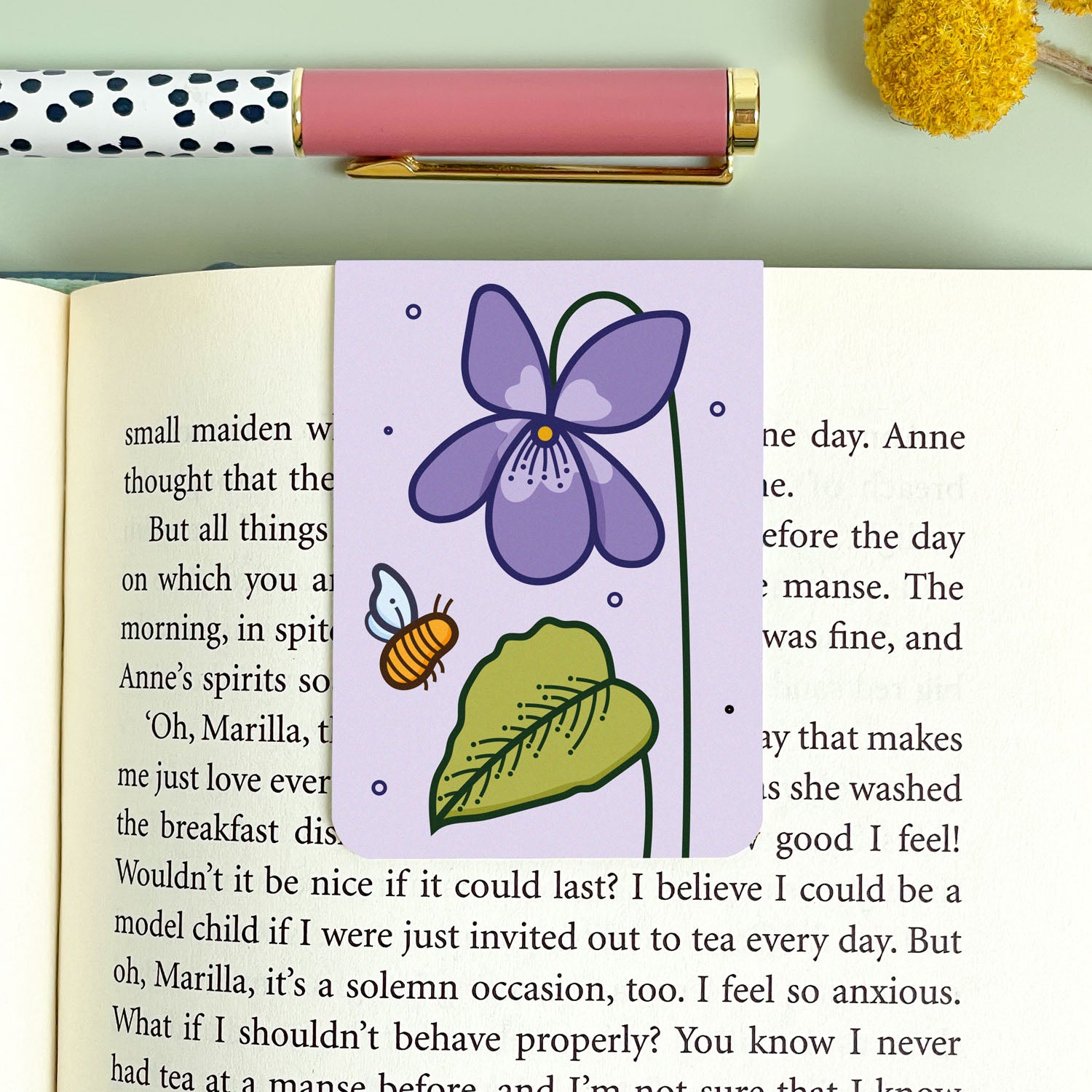 Floral violet bookmark with a purple flower, green leaf, and bee on an open book.