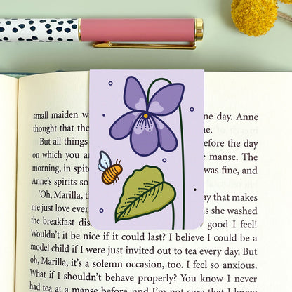 Floral violet bookmark with a purple flower, green leaf, and bee on an open book.