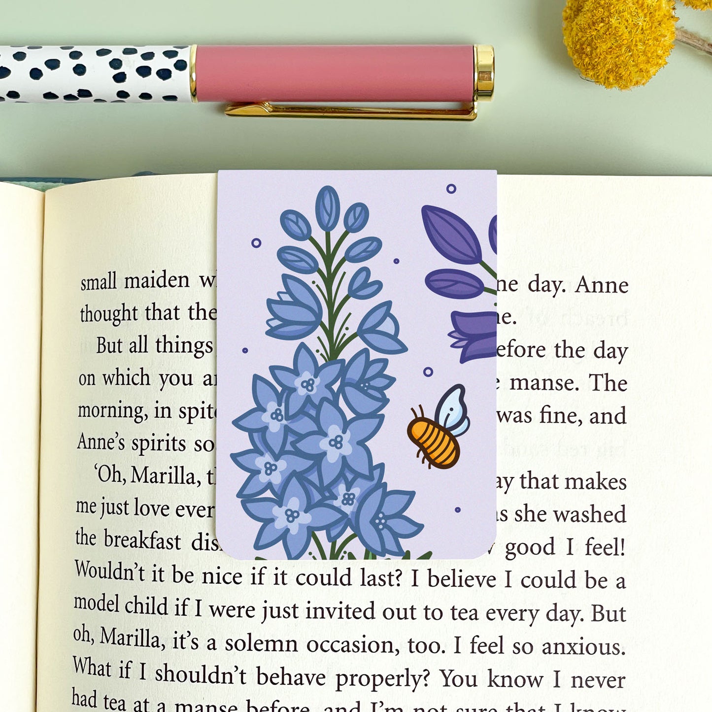 Purple floral bookmark with larkspur illustration and a bee on an open book, next to a polka dot pen and yellow flower.