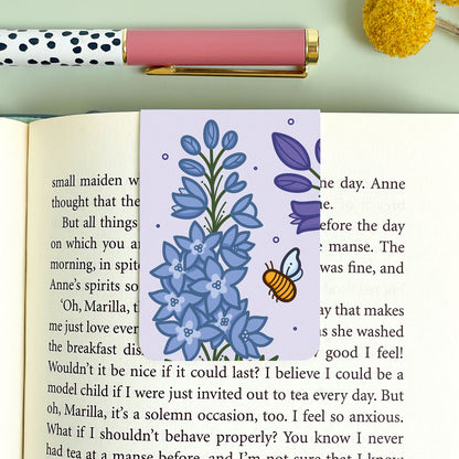 Purple floral bookmark with larkspur illustration and a bee on an open book, next to a polka dot pen and yellow flower.